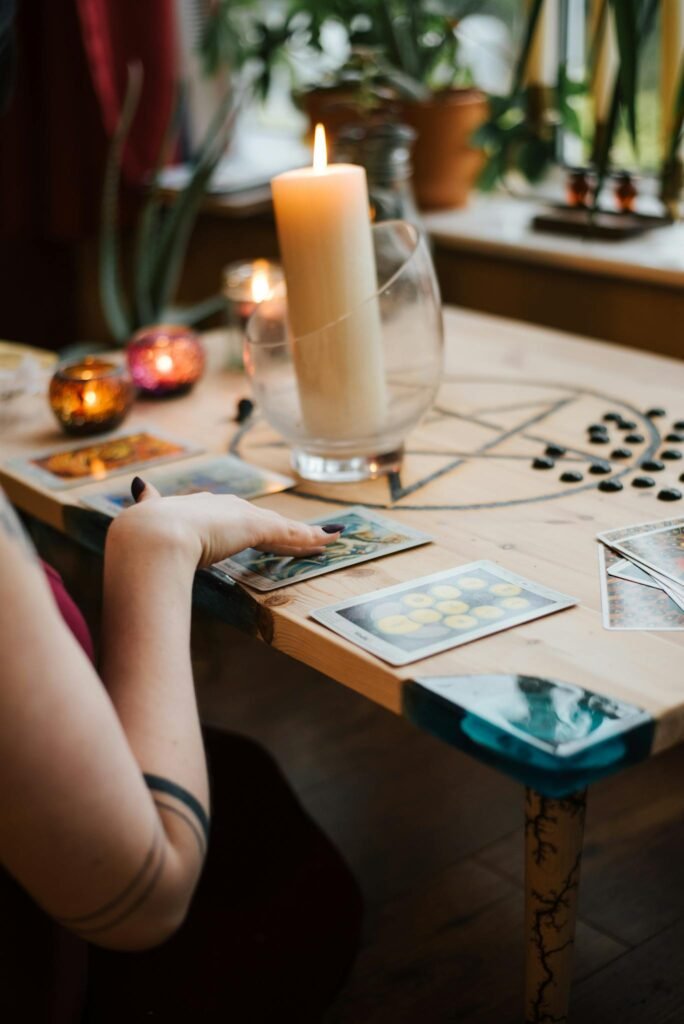 Crop unrecognizable female fortune teller predicting fate with tarot cards near burning candles on table at home